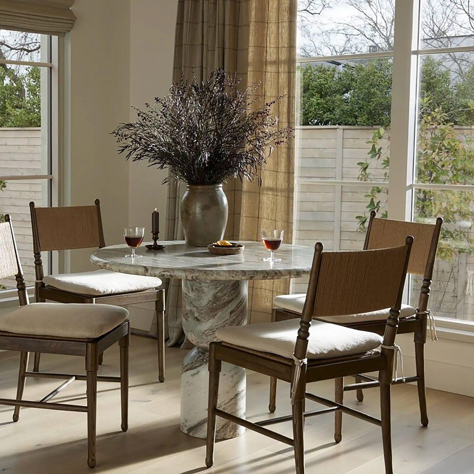 Dining set with four wooden chairs around a round marble table, near windows