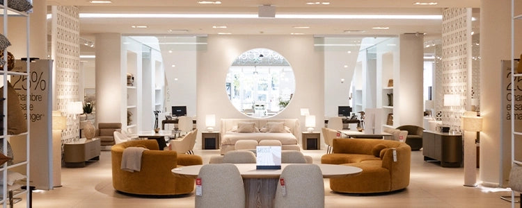 A wide interior showroom scene with multiple upholstered seating pieces, tables, lamps, and decorative screens arranged in a bright, modern furniture store. The central focus is a large round window above a sofa, with mustard yellow curved seating pieces in the foreground and neutral-toned furniture throughout the space.